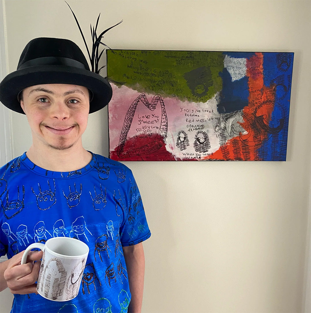 Artist Joel "J Positive" Jamensky standing in front of a beige wall with his colourful painting hung on it, while wearing a blue t-shirt and holding a white mug, both the t-shirt and mug have his art printed on them.