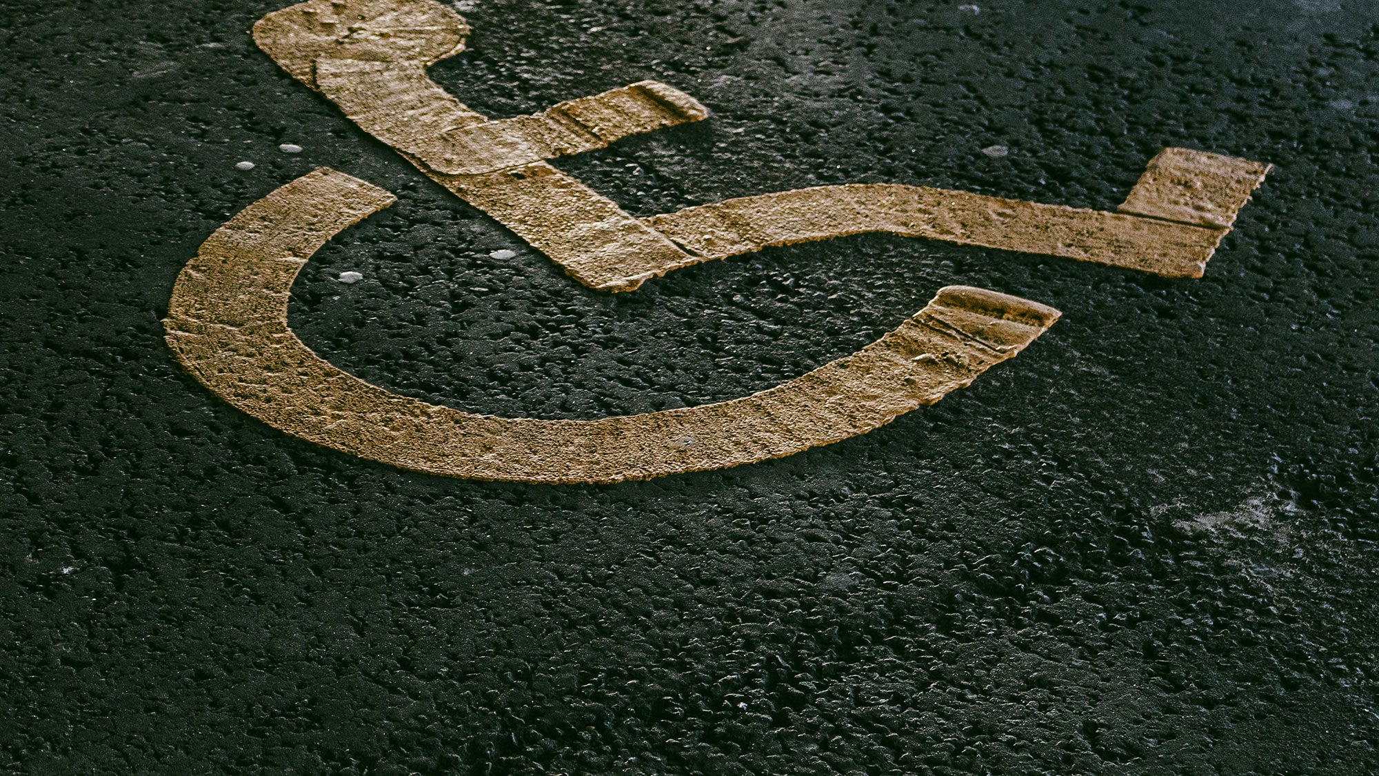 A moody, abstract photo of a symbol depicting a person in a wheelchair painted in yellow on concrete, denoting accessible parking