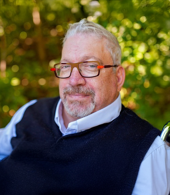 An upper-body photo of Max Brault sitting in a wheelchair outside in front of bright green foliage while wearing a light blue dress shirt under a navy blue knitted vest, and modern black full-rimmed glasses with red accents. He has blue eyes, has short silver hair and goatee. He looks directly at the camera with a slight and confident smile.